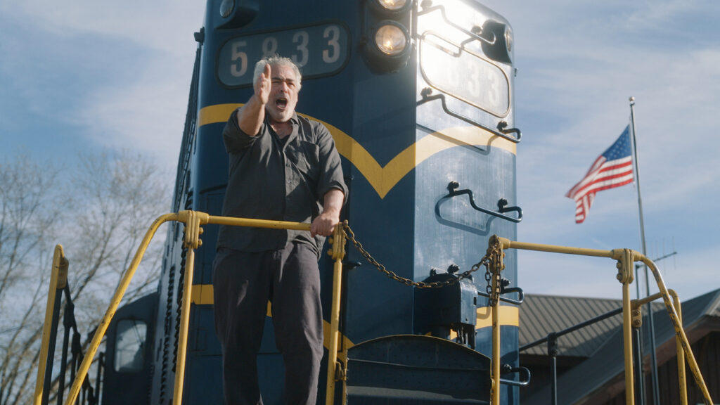 A man stands on the front platform of a blue and yellow locomotive, gesturing with his right hand. An American flag waves in the background under a clear sky.