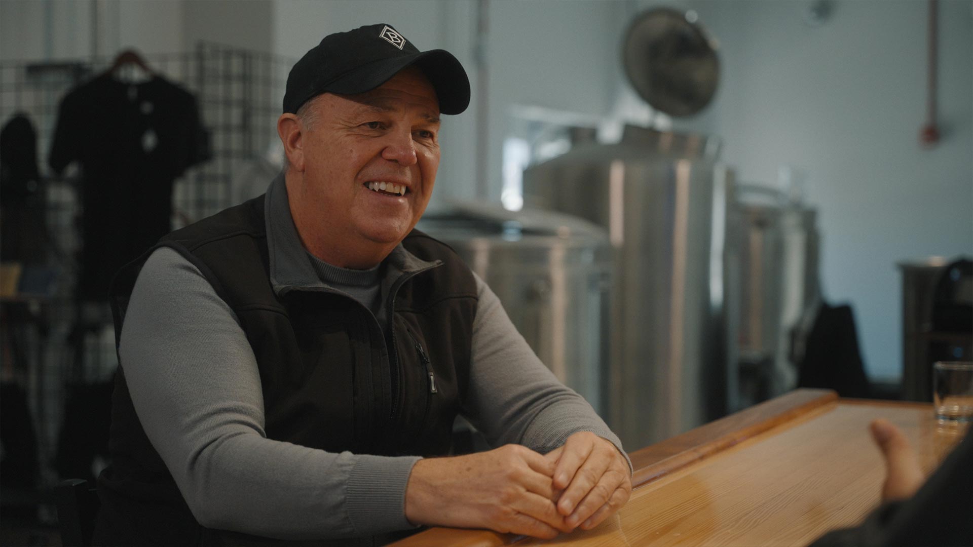 A smiling man in a black baseball cap, gray sweater, and black vest sits at the Market Bar inside Echo Spirits, a modern brewery with metal brewing tanks in the background.