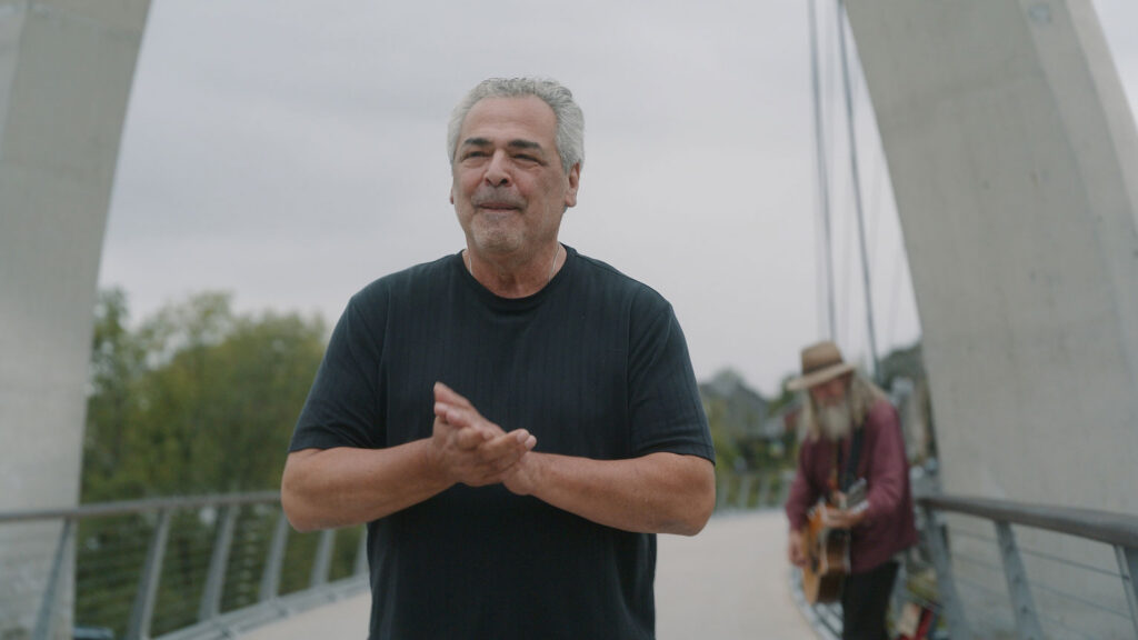 An older man in a black t-shirt stands on a modern bridge, clapping his hands. In the background, a person with long hair and a hat plays guitar. The sky is overcast and trees are visible.