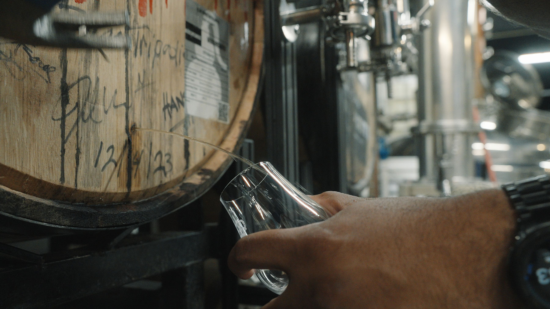 A close-up of a person’s hand holding a glass under a spout in a wooden barrel, collecting liquid. The barrel is marked with handwritten text and numbers. Metal pipes and brewing equipment are visible in the background.
