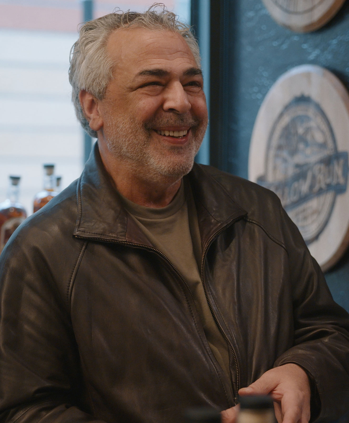 A smiling older man with gray hair and beard, wearing a brown leather jacket and olive shirt, stands indoors near round wooden wall plaques and bottles.