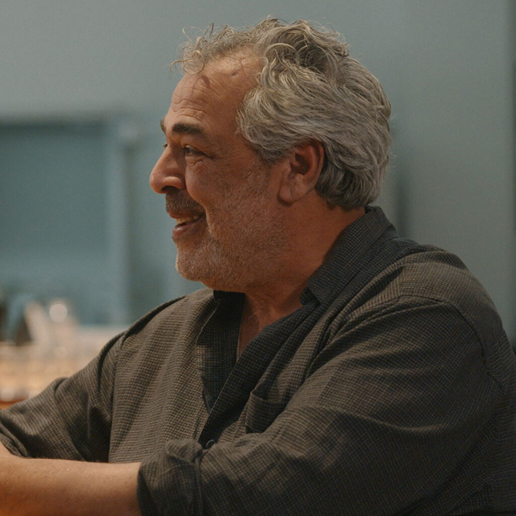 A middle-aged man with gray hair and a beard, wearing a dark button-up shirt, sits and smiles while looking to the left in a softly lit indoor setting.