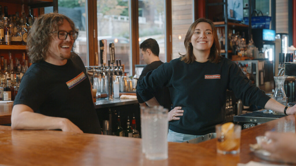 Two smiling bartenders stand behind a wooden bar in a well-lit pub, with drinks on the counter and bottles on shelves in the background. Both wear black shirts with name tags, and one person stands beside bar taps.