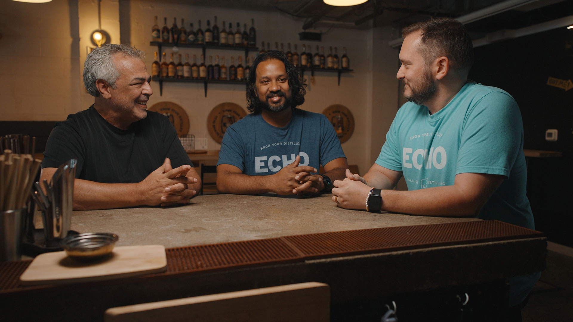 Three men sit at a bar counter in a cozy, dimly lit bar, smiling and talking. Two wear blue ECHO T-shirts, while the third wears black. Bottles and wooden barrels line the shelves behind them.