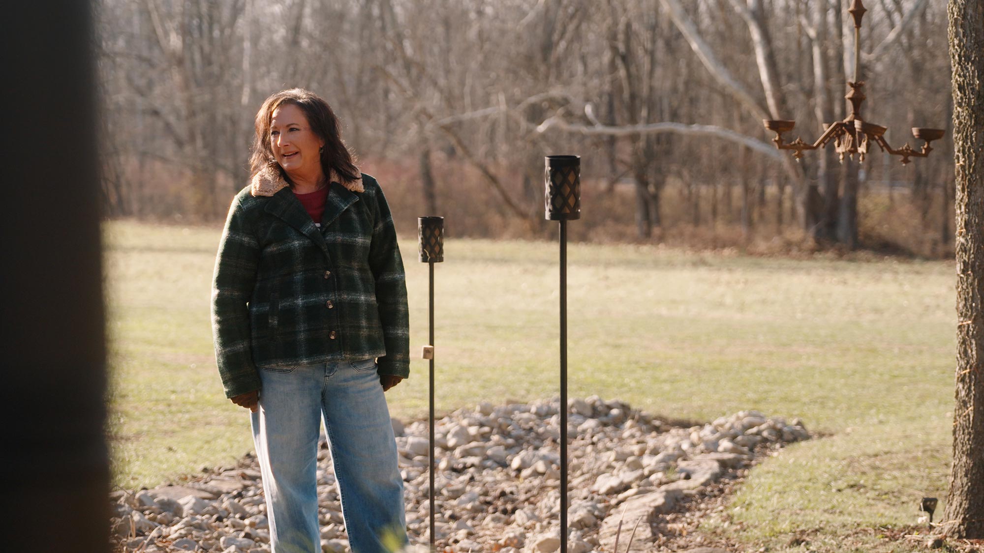 A woman in a plaid jacket and jeans stands outdoors on a grassy lawn near some rocks, metal garden torches, and bare trees—perhaps enjoying the crisp air outside Black Diamond Brewery & Distillery or The Mine Tavern.