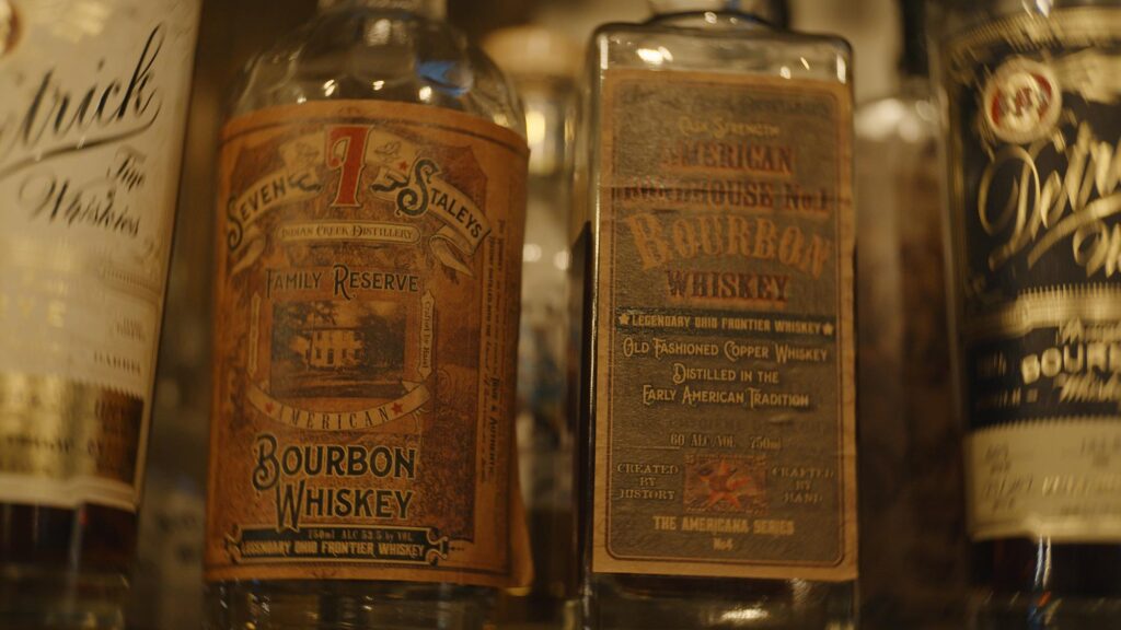 Close-up of several vintage bourbon whiskey bottles with ornate, detailed labels from brands like Black Diamond Brewery & Distillery, arranged side by side on a shelf at The Mine Tavern. The focus is on the central bottles and their aged designs.