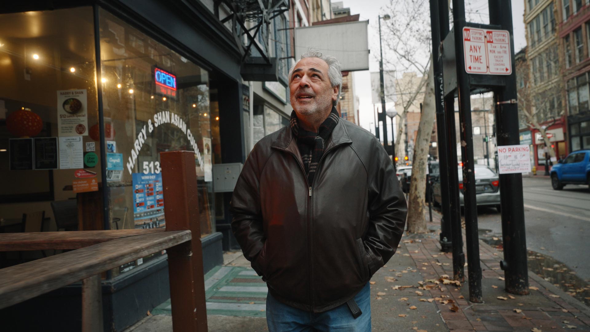 A man with gray hair and a beard, wearing a brown leather jacket and jeans, stands on a city sidewalk near Bay Horse Cafe, hands in his pockets and smiling. In the background, buildings advertise Willow Run Custom Bourbon.