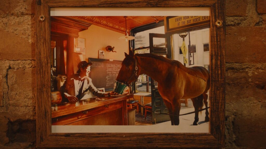 A framed photo shows a bartender serving a Willow Run Custom Bourbon to a horse standing at the bar inside Bay Horse Cafe, with warm lighting and rustic décor visible in the background.