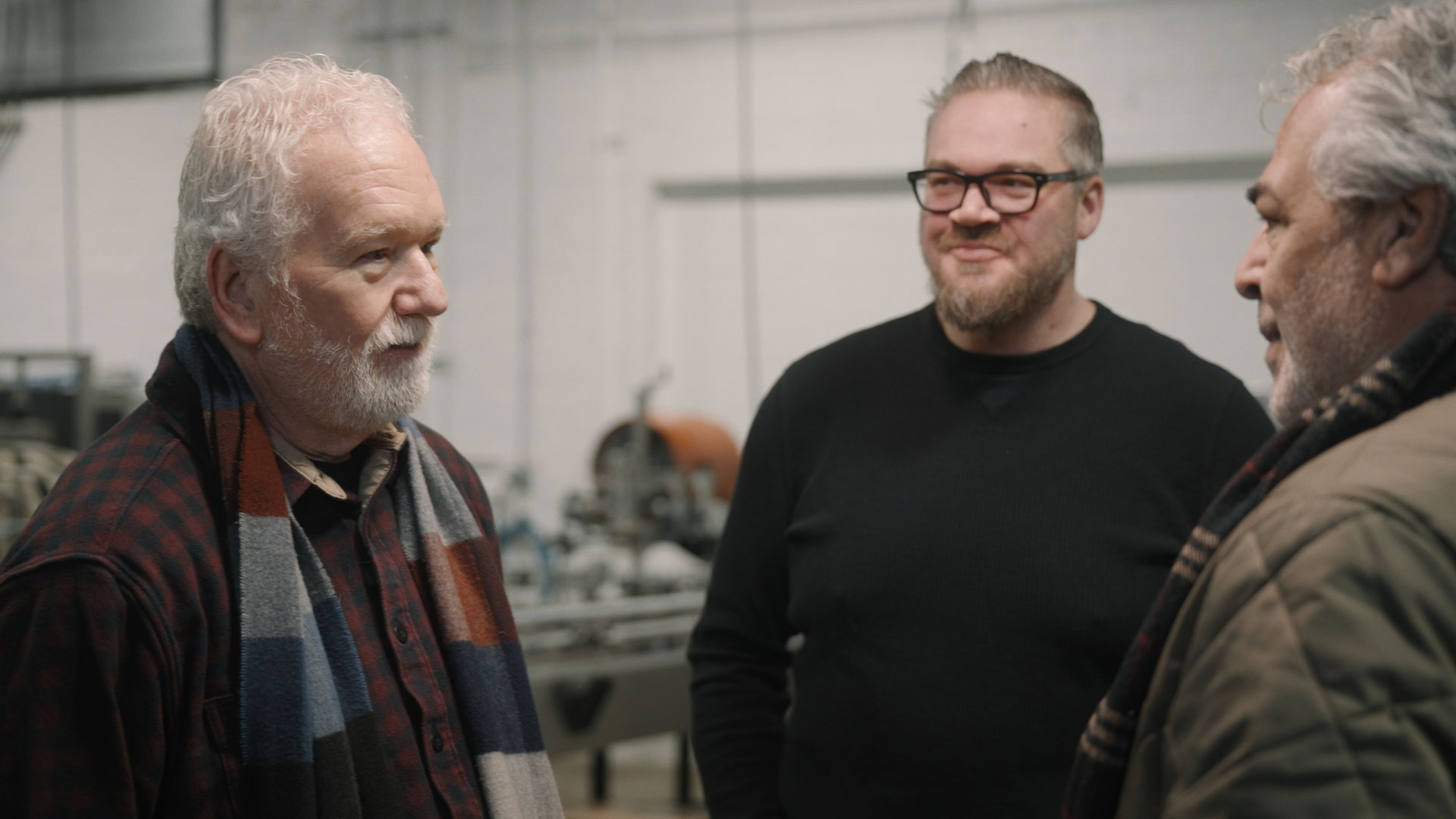 Three men stand indoors having a conversation at Western Reserve Distillers. One wears a plaid shirt and scarf, another sports glasses and a black sweater, while the third, in a brown jacket, stands near industrial equipment and Pizza DiLauro signage.