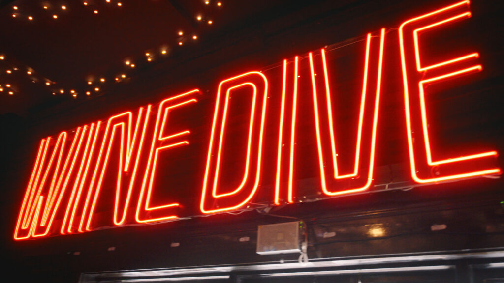 Bright red neon sign reading “WINE DIVE” glows against a dark background, with string lights above, creating a cozy, inviting atmosphere—perfect for sipping Western Reserve Distillers spirits or sharing a Pizza DiLauro.