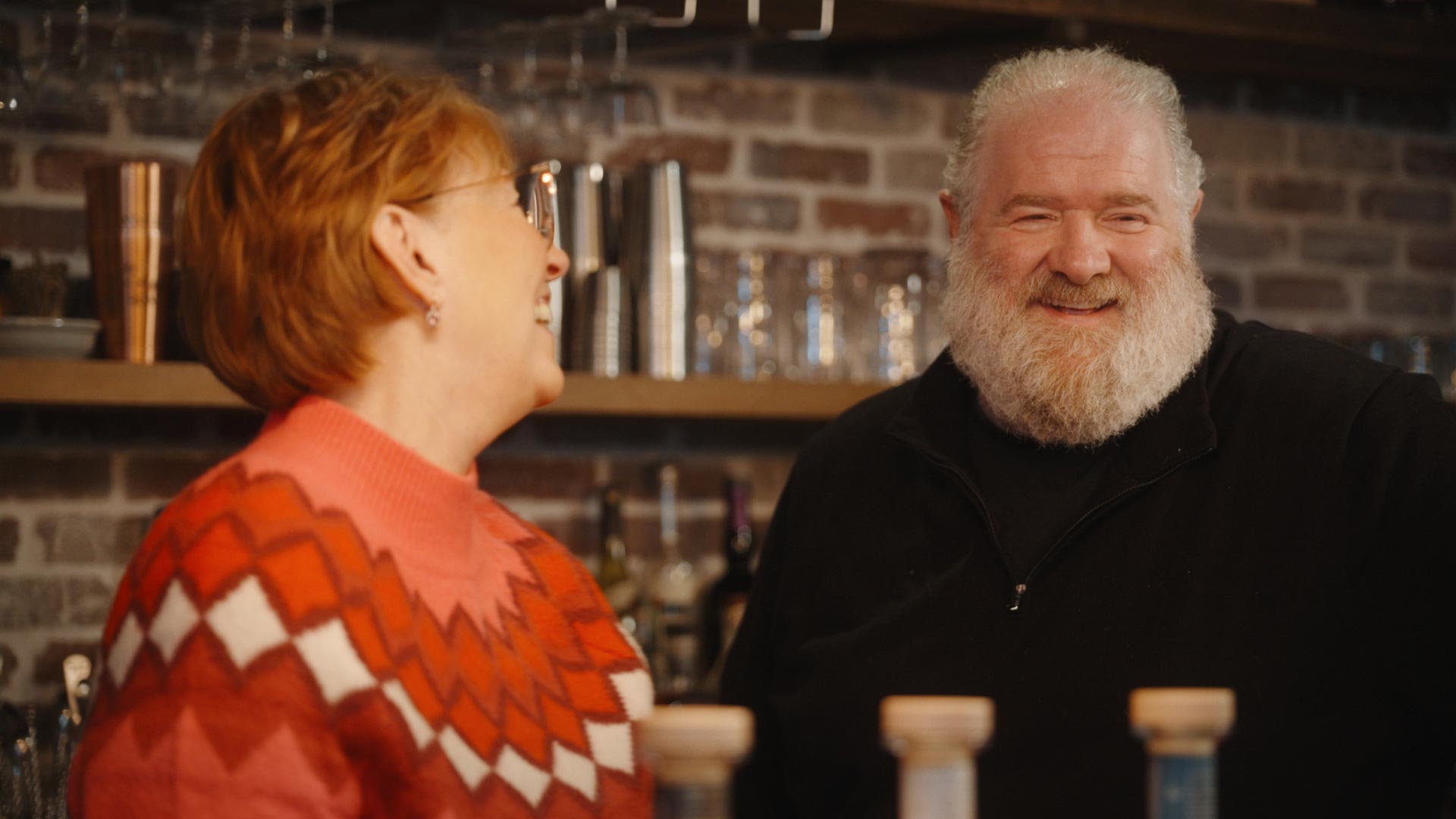 A woman in a red patterned sweater and a man with a white beard and black shirt smile and chat in front of shelves with glasses and bottles from Western Reserve Distillers, set against a brick wall, perhaps discussing their favorite Pizza DiLauro.