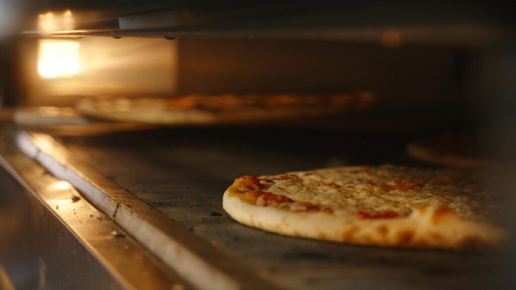 Close-up of Pizza DiLauro pizzas baking inside an oven, with a warm light illuminating the cheese and crust as they cook on the metal baking surface at Western Reserve Distillers.
