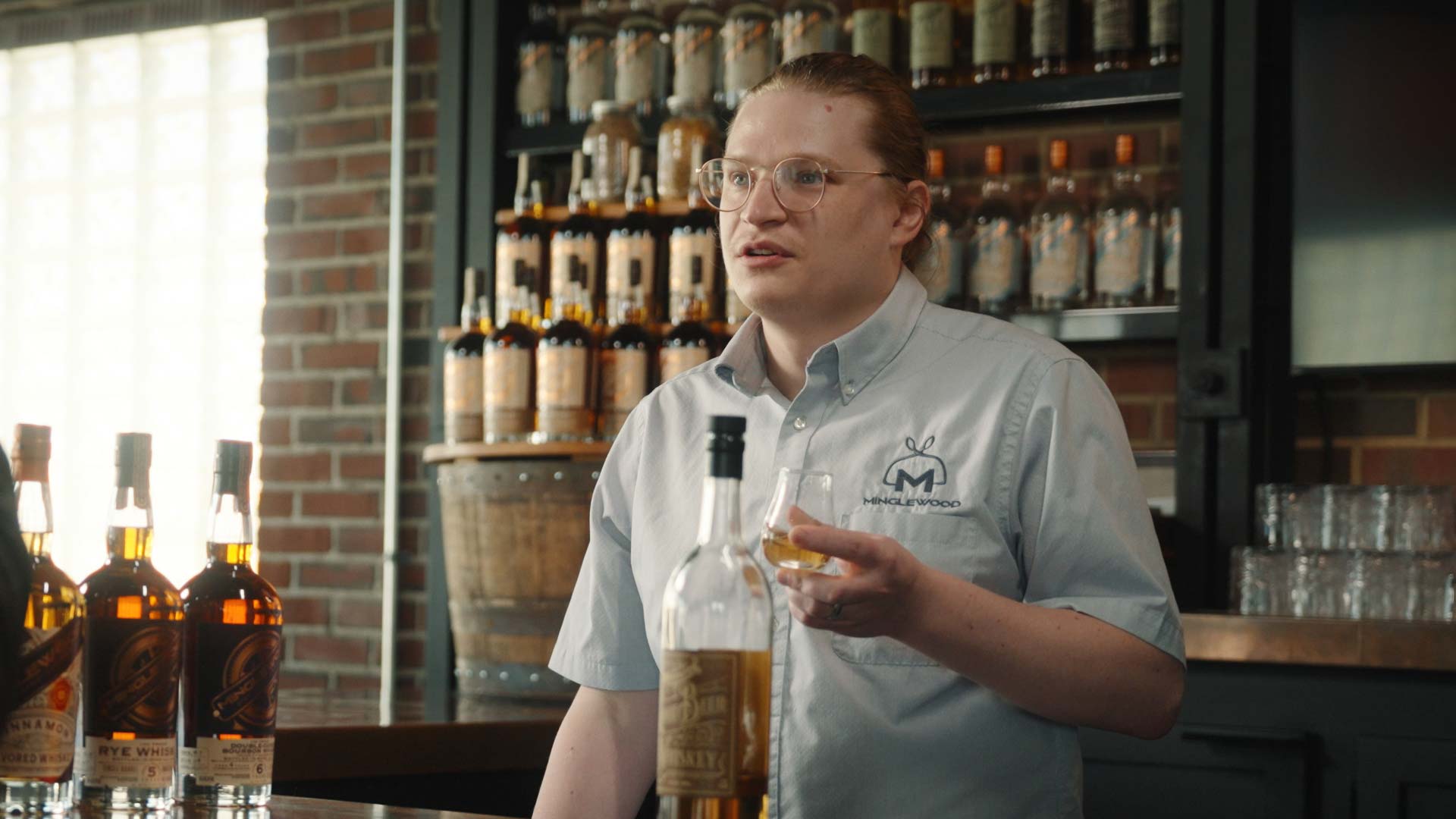 A person with glasses and a light blue shirt stands behind a bar at High Bank Distillery Co., holding a glass of whiskey. Several bottles line the counter, framed by shelves in the background, creating a good land for whiskey lovers.