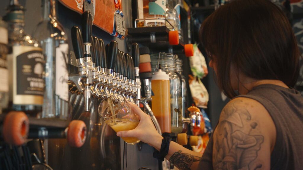 A person with a visible arm tattoo pours High Bank Distillery Co. beer from a tap into a glass at a lively bar, surrounded by bottles and Good Land drink taps, creating a casual and energetic atmosphere.