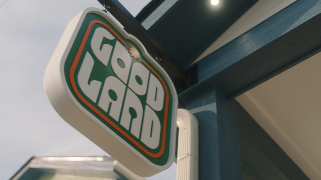 A rectangular sign reading GOOD LAND in bold, retro-style white letters with green and orange outlines hangs outside a blue building near Cleveland Whiskey + Wine Dive.