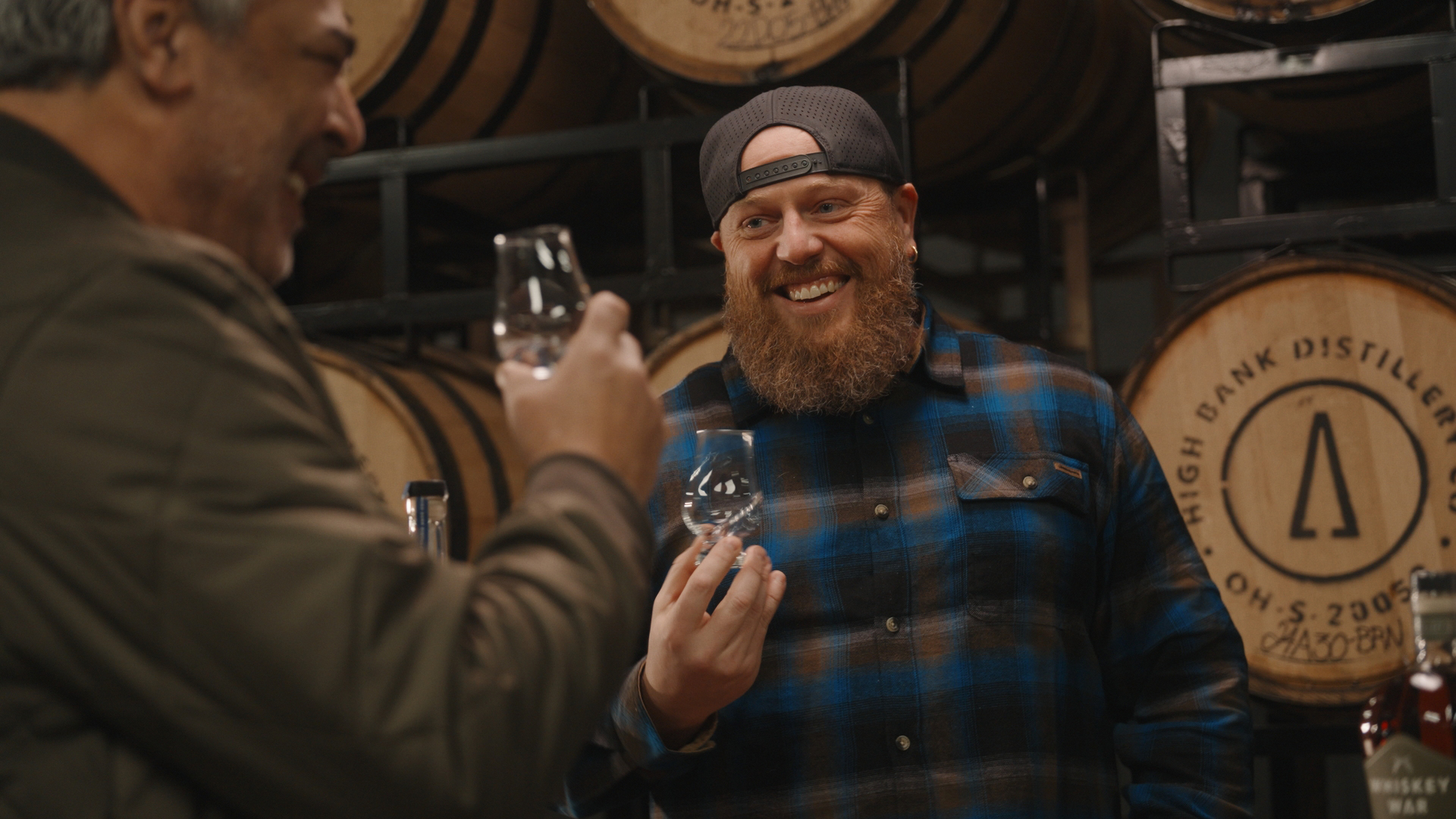 Two men enjoy a conversation while holding Cleveland Whiskey glasses in front of stacked barrels at a distillery. One, wearing a flannel shirt and backward cap, smiles warmly—capturing the relaxed spirit of Wine Dive gatherings.
