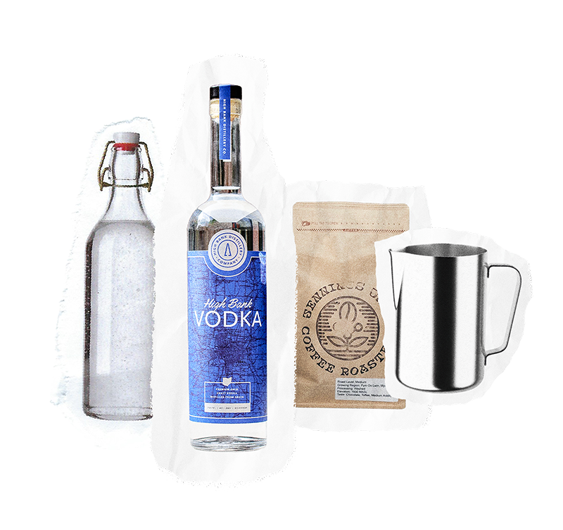 A clear glass bottle, a tall bottle of vodka, Cleveland Whiskey, a brown bag of coffee beans, and a metal pitcher are arranged in a row on a white background at Wine Dive.