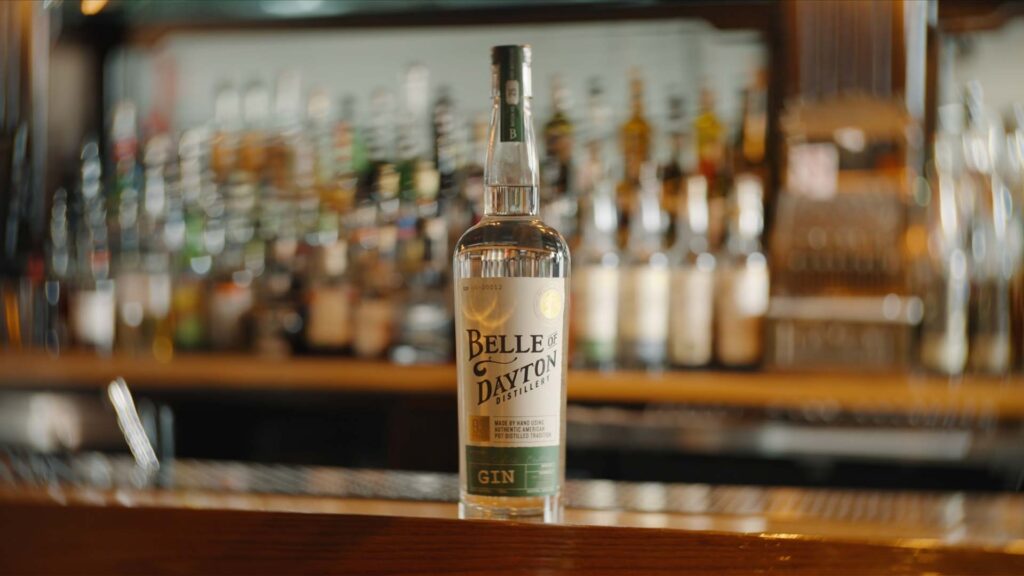 A bottle of Belle of Dayton Distillery gin sits on a wooden bar counter at Crafted & Cured, with blurred bottles and bar equipment in the background.