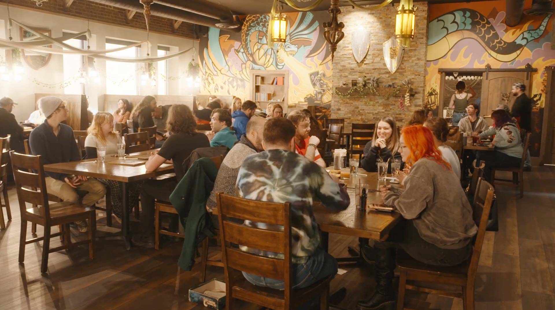 A cozy, colorful cafe with mural-covered walls evokes the charm of The Green Dragon Inn. People sit at wooden tables, chatting and enjoying drinks from Towpath Distillery, all beneath warm lighting and rustic decor in a lively atmosphere.