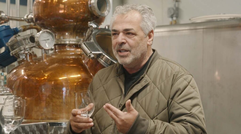 A man with gray hair and a beard, wearing a quilted jacket, holds an empty glass while talking in front of Towpath Distillery’s large copper distillation apparatus, evoking the spirit of The Green Dragon Inn in this industrial setting.